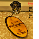 Ovale Rindenscheibe mit schwarzer Gravur die lautet Herzlich Willkommen Steelamore, in der oberen rechten Ecke das Logo von Steelamore , auf der Rindenscheibe steht senkrecht eine Rose aus Edelstahl mit silbernen Rosenblütenblättern und silbernem Stiel sowie silbernen Blättern